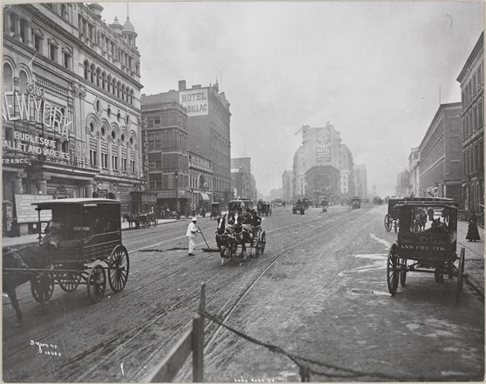 A black-and-white photograph showing the intersection of Broadway and 42nd Street, the present-day location of Times Square. The city street is surrounded by multistory buildings, and horse-drawn carriages drive in the street. In the center of the street is a New York City Sanitation worker dressed in white.